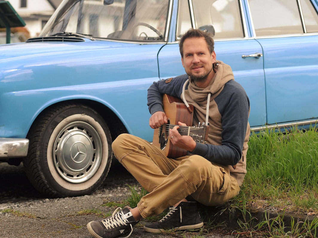 Mike Kremer von Miljö spielt sitzend Gitarre vor einem Auto