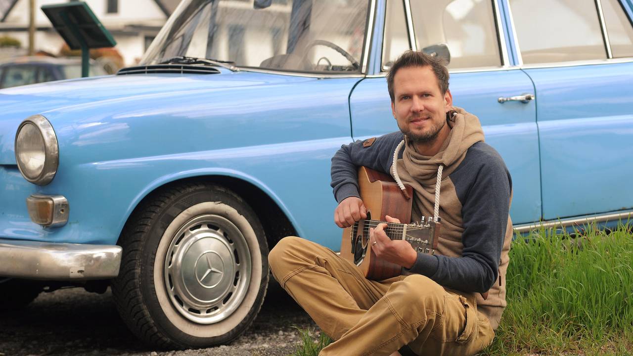 Mike Kremer von Miljö spielt sitzend Gitarre vor einem Auto